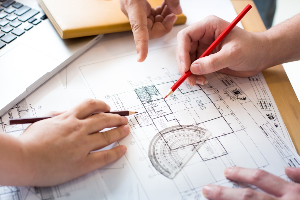 In den Projektateliers werden architektonische und städtebauliche Fragestellungen als Entwurfsaufgaben bearbeitet – in Gruppen von höchstens 25 Personen. Hände mehrerer Personen über einem ausgebreiteten Bauplan auf einem Besprechungstisch