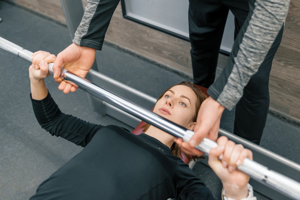 Die Fitnesstrainer B-Lizenz ist bei der Deutschen Sportakademie der Einstieg in die Trainerarbeit auf der Fläche. Fitnesstrainer sichert eine junge Frau beim Bankdrücken im Fitnessstudio