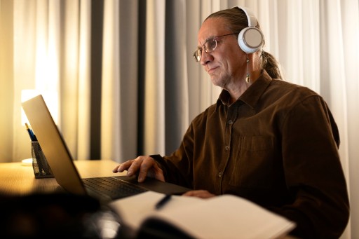 Die Frage ist selten, ob KI erlaubt ist. Die Frage ist, ob du am Ende jeden Satz erklären kannst. Studierender mit Kopfhörern arbeitet abends am Laptop, daneben ein aufgeschlagenes Notizbuch