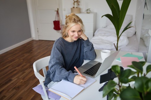 Junge Frau sitzt an ihrem Schreibtisch in der Wohnung vor Laptop und Notizheft