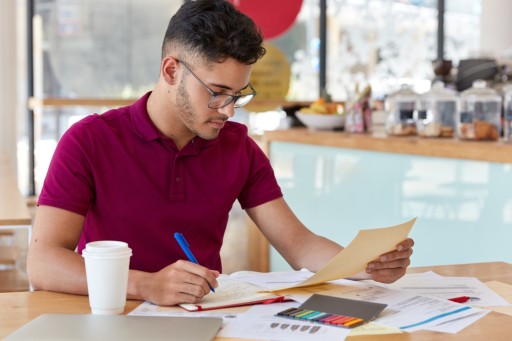 Erst wenn klar ist, welche Textart verlangt wird, lässt sich der Aufwand seriös schätzen. Studierender sichtet an einem Cafétisch ausgedruckte Unterlagen und macht sich handschriftliche Notizen