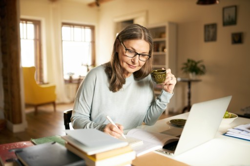 Das Fernstudium an der IU läuft vollständig online, Lernzeiten legen Studierende selbst fest. Frau arbeitet in einem hellen Raum am Laptop, daneben Bücher und ein Notizbuch